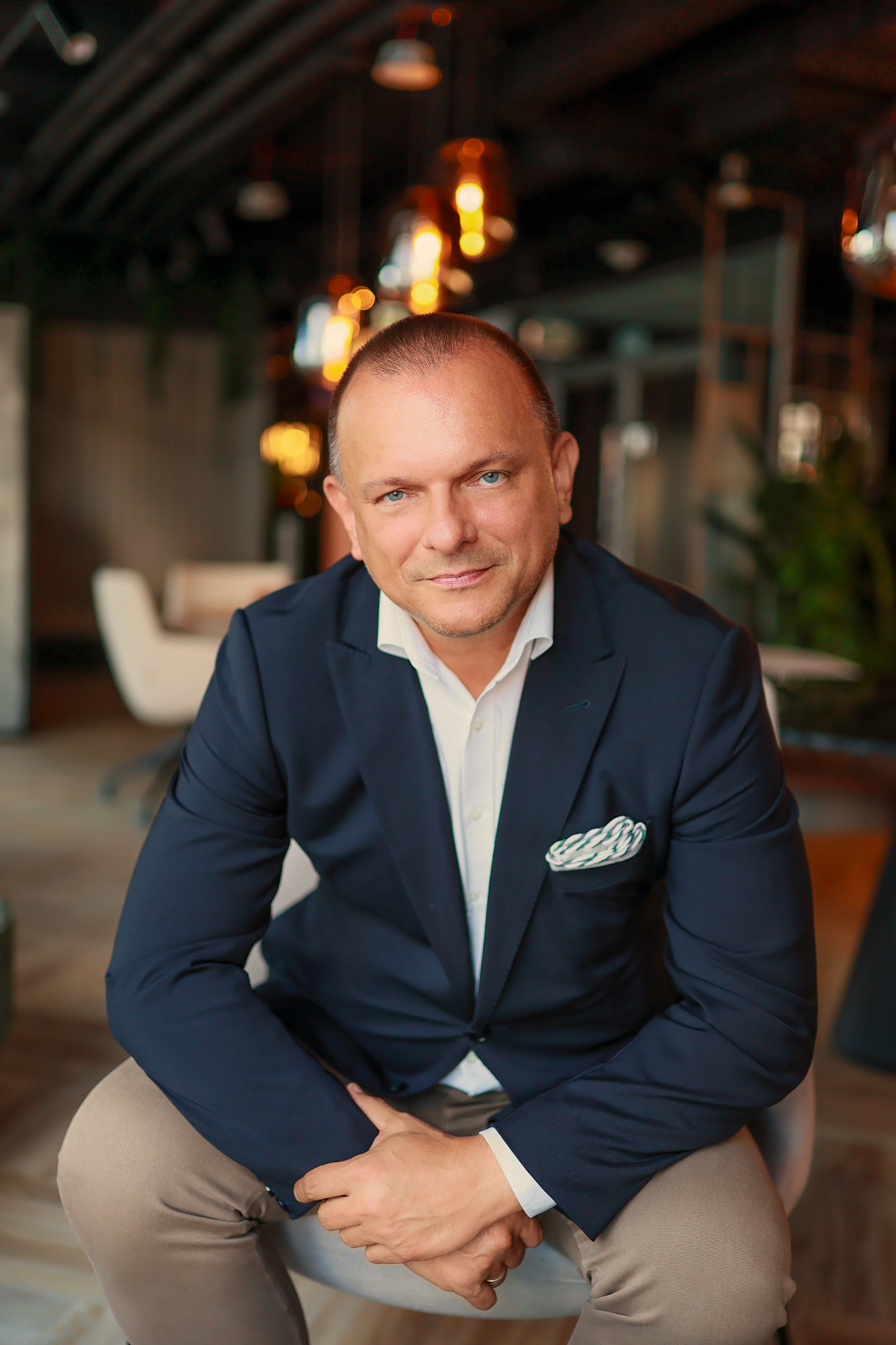 Portret uśmiechniętego mężczyzny w eleganckim stroju, stojącego w jasnym pomieszczeniu z elementami biura w tle.